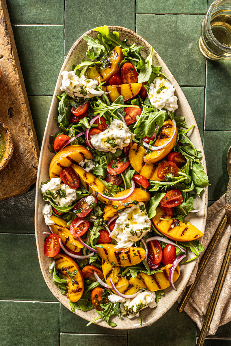 arugula salad with grilled peaches burrata and tomatoes on green tiles backdrop